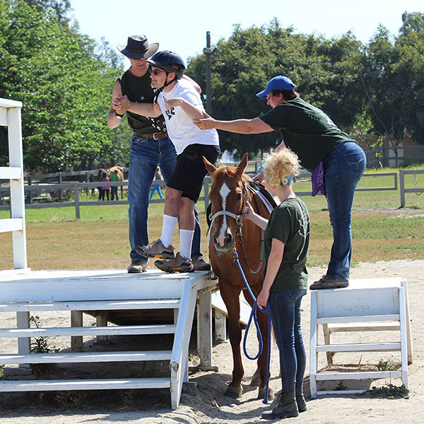Two experienced Ivey Ranch volunteers assist a therapeutic riding participant from a mounting block onto a chestnut horse, with a third volunteer holding the horse steady at the arena.