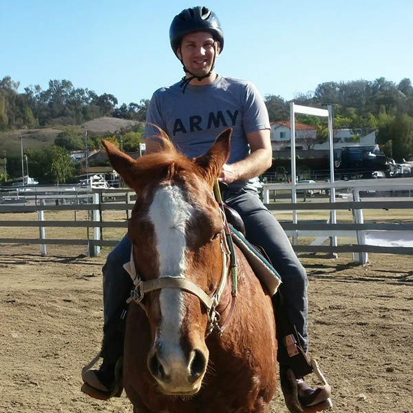 A smiling Army veteran in a helmet rides a chestnut horse in the arena during the Horses for Heroes program at Ivey Ranch in Oceanside, CA