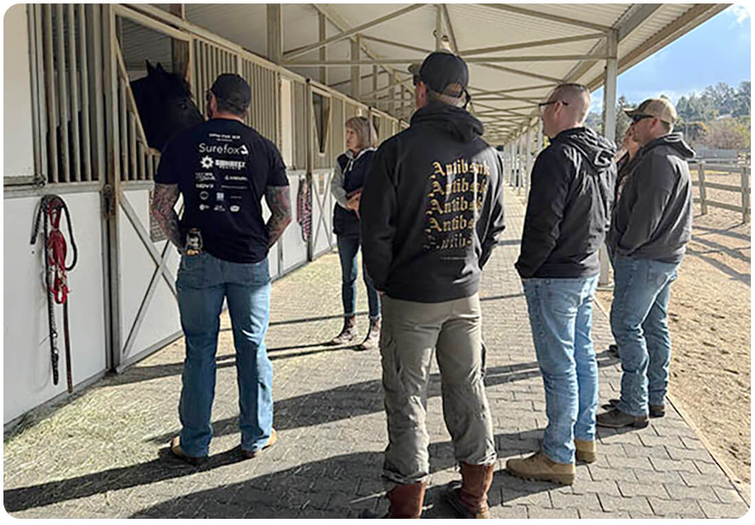 A group of veterans gathers along the stable walkway with a black horse during the Horses for Heroes program at Ivey Ranch in Oceanside, CA