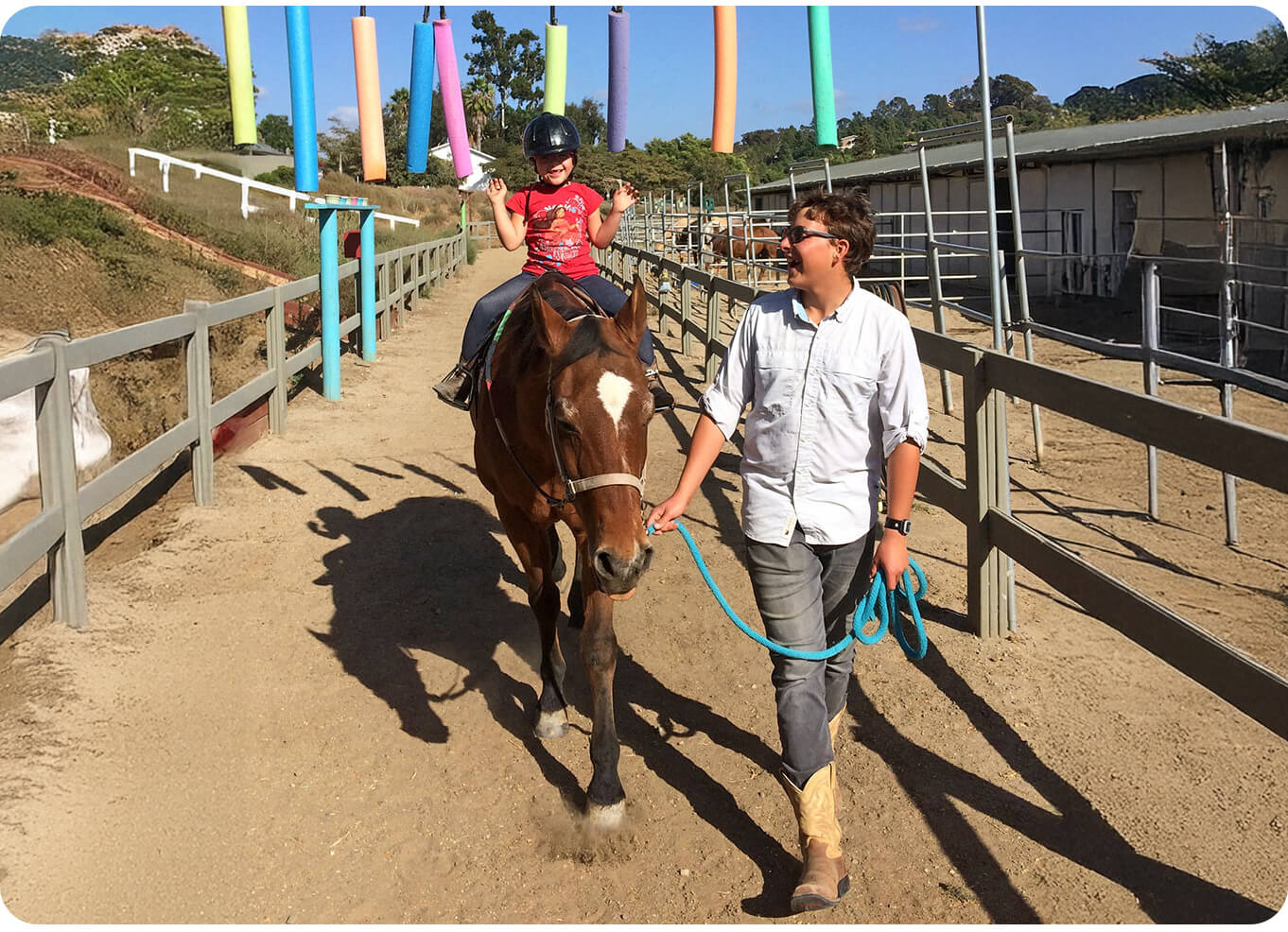 A smiling child in a helmet rides a chestnut horse with arms raised while a horse leader guides them through the arena at Ivey Ranch in Oceanside, CA