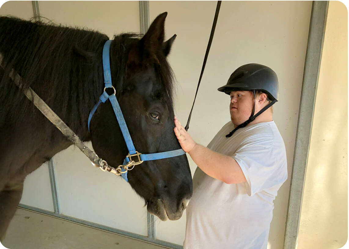 A young adult in a riding helmet gently touches the face of a black horse in a stable at Ivey Ranch in Oceanside, CA