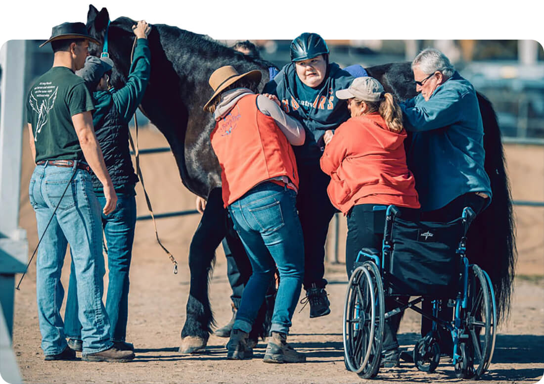 Volunteers and sidewalkers assist a rider in a helmet onto a black horse during a Therapeutic Horseback Riding session at Ivey Ranch in Oceanside, CA
