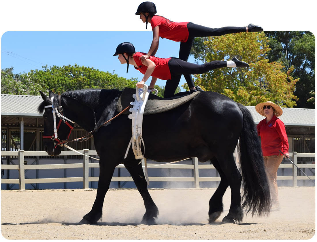 Two vaulters in red uniforms perform a stacked pose on a moving black horse while an instructor guides from the ground at Ivey Ranch in Oceanside, CA