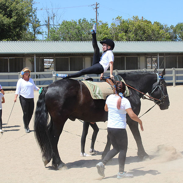 A young vaulter performs a split on a dark horse while instructors guide from the ground during a vaulting session at Ivey Ranch in Oceanside, CA