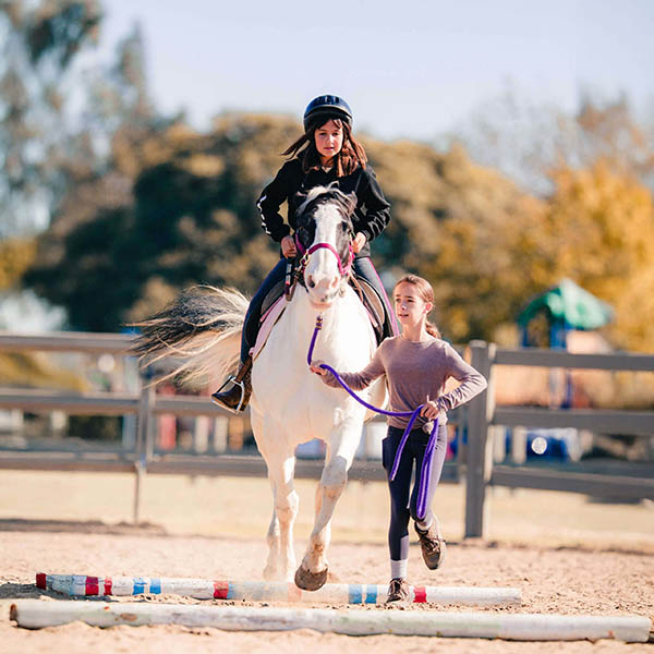 A young rider on a white pinto horse cantering over ground poles with a leader during an able-bodied riding lesson at Ivey Ranch in Oceanside, CA
